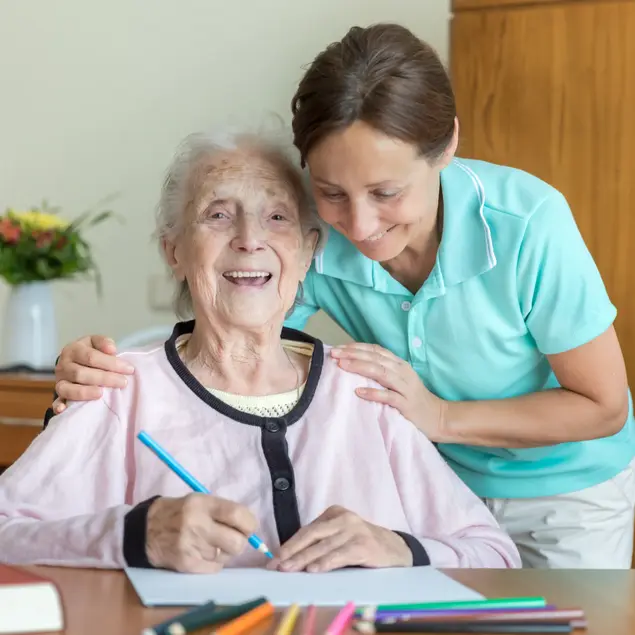 Eine Pflegerin beugt sich zu einer älteren Frau und berührt sie an den Schultern. Die ältere Frau lacht und hält einen Stift in der Hand.