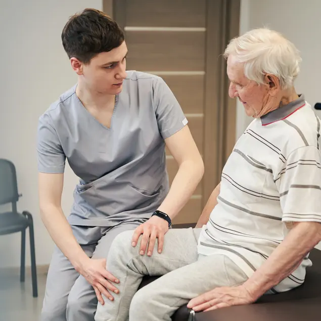 Ein junger Physiotherapeut therapiert das Bein eines älteren Mannes, der auf einer Behandlungsliege sitzt.